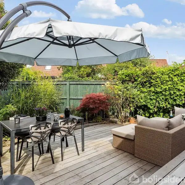 Stor hvid parasol står åben over havemøbler på en træterrasse. Bord og stole i sort metal står til venstre, mens sofa i rattan står til højre. Grønne buske omgiver terrassen.