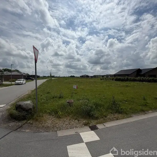 En vejskilt står ved siden af en græsmark i et boligområde med flere huse. En parkeret bil ses på vejen. Skyet himmel danner baggrunden. Et lille skilt står på marken.
