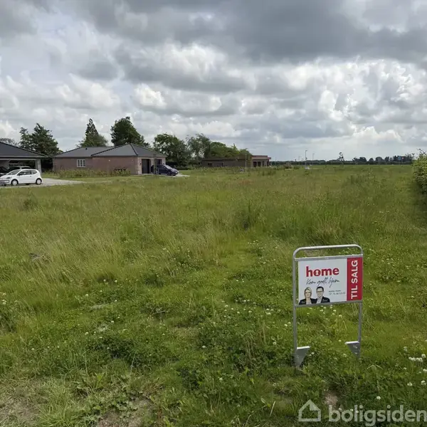 Græsmark med skilt, der siger "home, kom godt hjem, TIL SALG", i forgrunden. Baggrunden viser en række huse og biler under en overcast himmel.