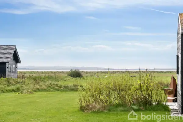 Et træhus står stille i en grøn have med udsigt over en strand og havet i baggrunden. Skyfri himmel og spredte buske omgiver stedet.
