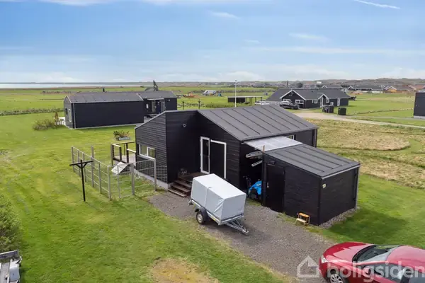 Sort træhus står stille på en stor grøn græsplæne. Husets terrasse er omgivet af et hegn, og en lys trailer er parkeret foran. Flere lignende huse ses i baggrunden.
