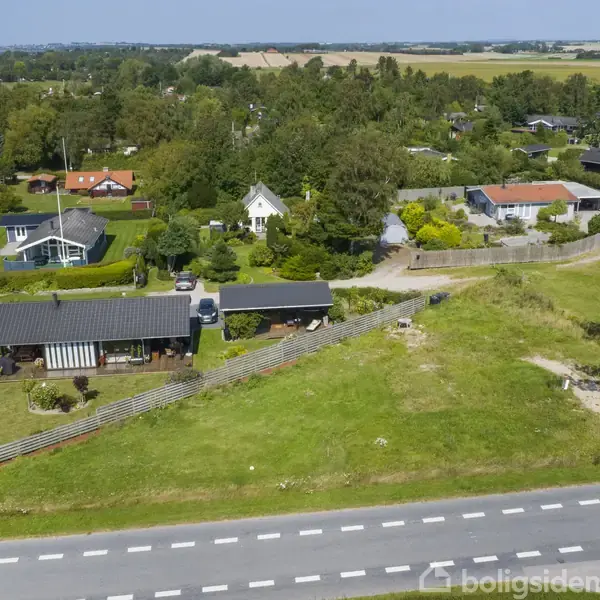 Hus på en grøn grund omgivet af andre huse i et landligt område. Foran løber en vej med hvide striber. Landskabet er frodigt med træer og marker i baggrunden.