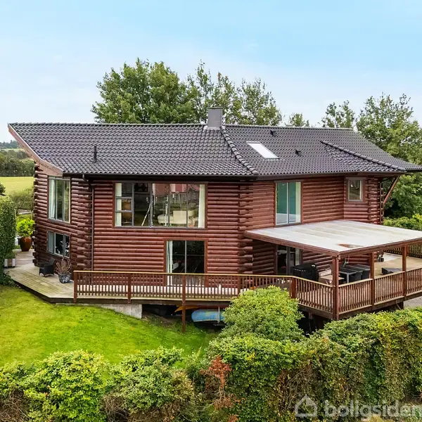 Et rummeligt træhus står blandt frodige hække og græsplæner. Det har store vinduer og en stor terrasse med tag, omgivet af landlig natur og nabohuse i baggrunden.