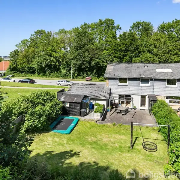 Et rækkehus med en græsplæne foran, hvor en trampolin og en gynge er placeret. Omkring huset er der buske og træer, samt en vej og andre huse i baggrunden.