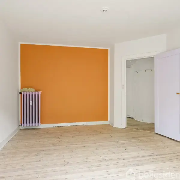 Et rum med en orange væg, hvor radiatoren er placeret foran. Døren til højre står åben, hvilket fører til en hvid gang. Gulvet er lyst træ.