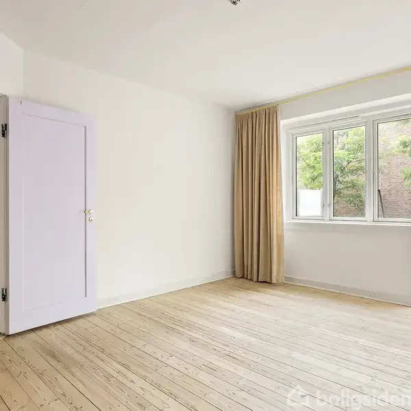 Et lyst rum med trægulv har en åben dør til venstre og store vinduer til højre med beige gardiner. Udenfor er der grønne træer. Ingen tekst til stede.