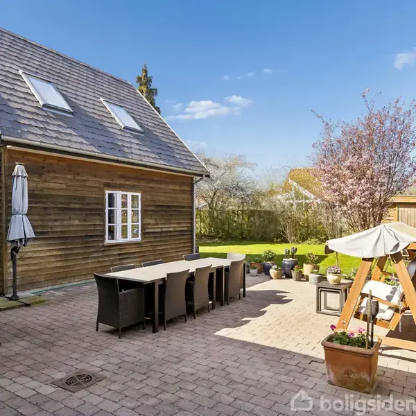 Et træhus med skråt tag står ved en stor terrasse med et langt spisebord og stole. I baggrunden ses en grøn græsplæne, blomstrende træer og et skur.
