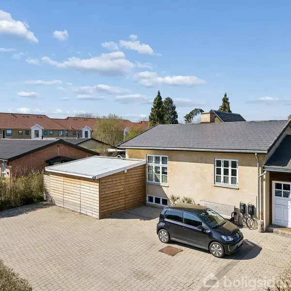 Bil parkeret på flisebelagt indkørsel foran et lysegult hus med sort tag. Huset har en tilknyttet carport og omgives af andre bygninger og træer på en solrig dag.