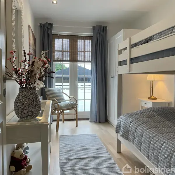 En seng med gråstribet sengetøj står i et lyst værelse. En vase med blomster står på et bord. Gardiner dækker en dør til en balkon. En lampe lyser op ved siden af sengen.