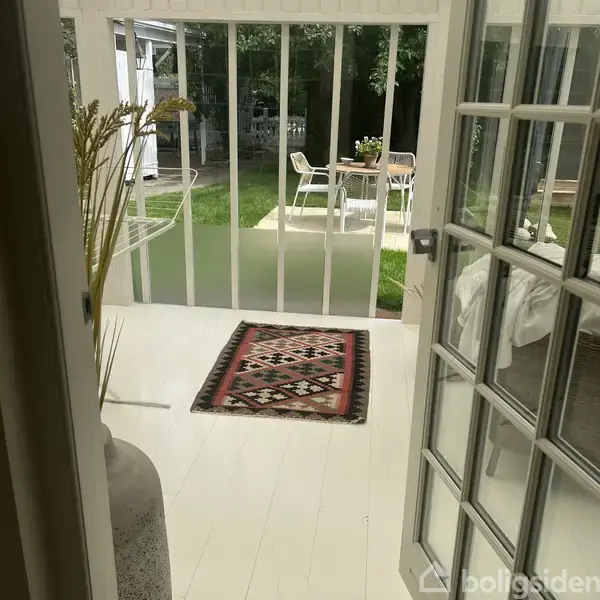 En dør åbner mod en terrasse med et tæppe på gulvet. Udenfor ses et havebord med stole og en grøn græsplæne i baggrunden.