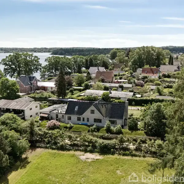 Hus omgivet af grønne haver ligger i et beboelseskvarter. I baggrunden ses en sø og skovomgivelser. Stille, frodig atmosfære omfatter flere parcelhuse og træer.