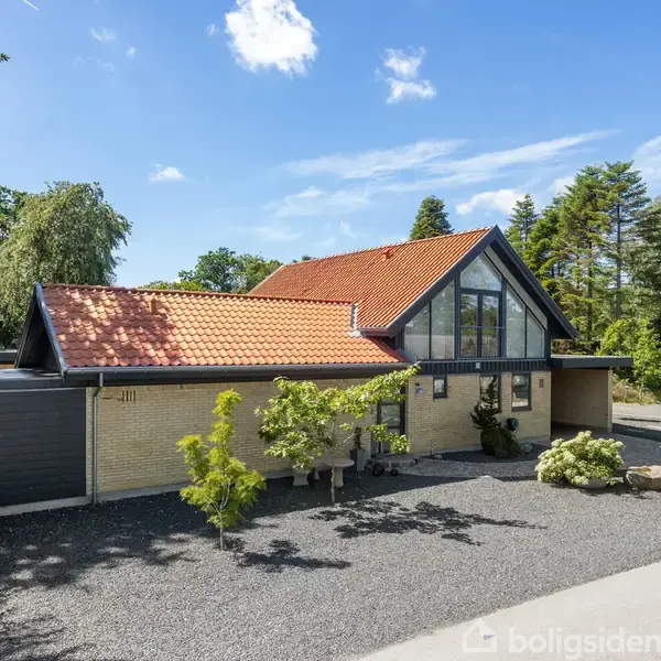 Et moderne hus med rødt tegltag og store glaspartier, omkranset af træer. Foran huset er en grusbelagt gårdsplads med dekorative planter. Skulpturer pryder indgangen, mens en bil er parkeret til højre.