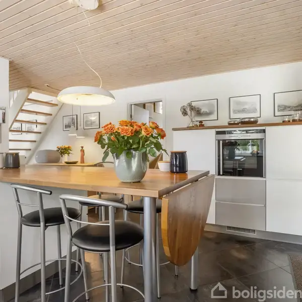 Køkken med hvid indretning og træloft. En ø med barstole og en vase med orange blomster dominerer rummet. Væghylde med billeder og en indbygget ovn i baggrunden.