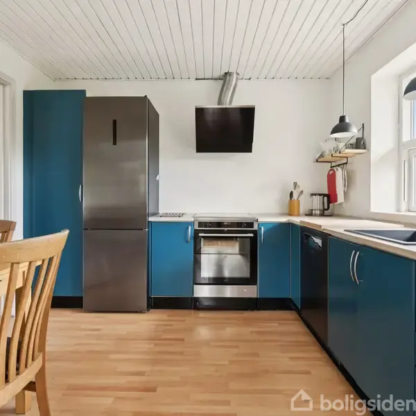 Køkken med blå skabe står i et lyst rum. Et køleskab og komfur er på plads ved væggen. Bord og stol er i træ. En stor vindue giver udsigt til grønne omgivelser.