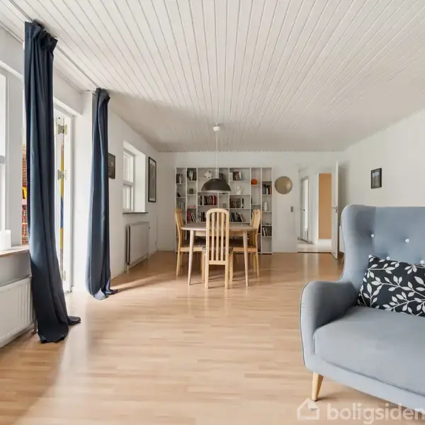 En lys stue med en blå lænestol i forgrunden. Et spisebord med stole er i baggrunden. Store vinduer med mørke gardiner giver udsigt til udendørsområder.