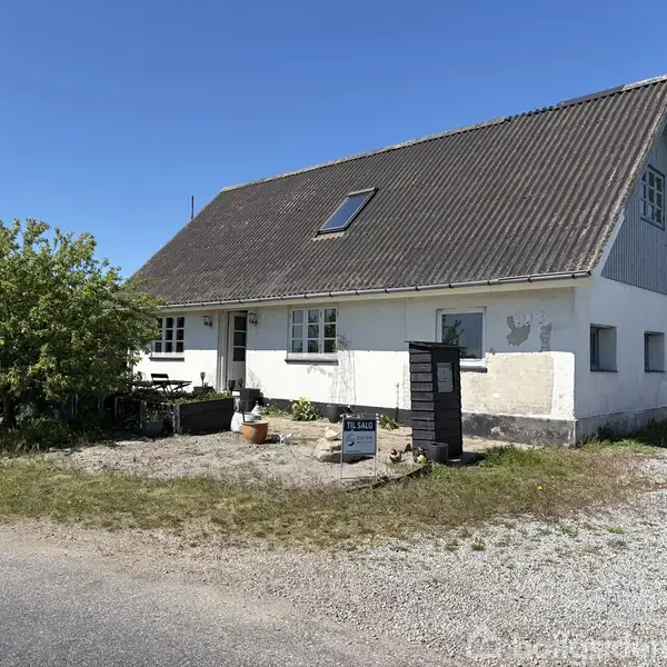 Hvidt, traditionelt hus med skråt tag står på en grusvej. En lyse dekoration findes på muren ved siden af hoveddøren. Foran huset står et skilt med teksten: "Til salg."