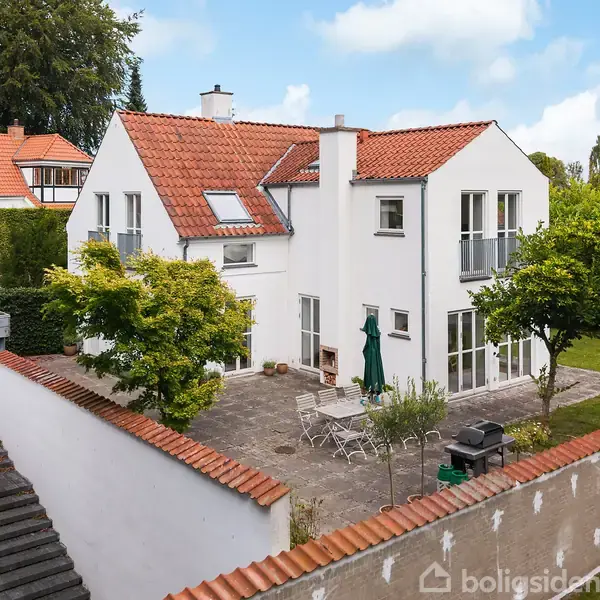 Et hvidt hus med rødt tegltag står i en grøn have. Foran huset er en terrasse med havemøbler og en parasol.