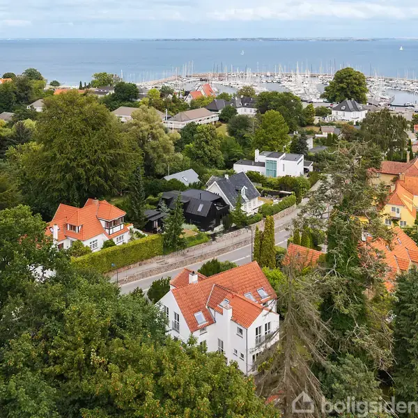 Huse med røde tage står blandt træer. I baggrunden ses en lystbådehavn ved kysten med mange både. Området er frodigt og grønt.