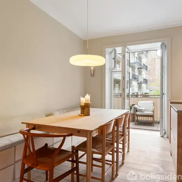 Et træbord står i en spisestue med seks stole, omgivet af en lys væg. Der er en pendellampe over bordet. Glasdøre fører ud til en terrasse med udsigt til bygninger.