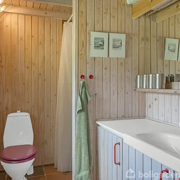 Et badeværelse med en hvid toilet med rødbrun sæde, en håndvask og spejl til højre. Rummet har træpaneler og små billeder på væggen. Der hænger et grønt håndklæde ved bruseren.