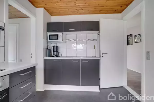 Et moderne køkken med grå skabe indeholder en mikroovn og kaffemaskine på en hvid flisevæg. Træloftet og åbne døre leder til en tilstødende stue.