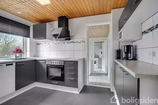 Et moderne køkken har grå skabe og en sort ovn. En kaffemaskine står på bordet. Gennem døren ses en vaskemaskine. Røde blomster er i vindueskarmen.