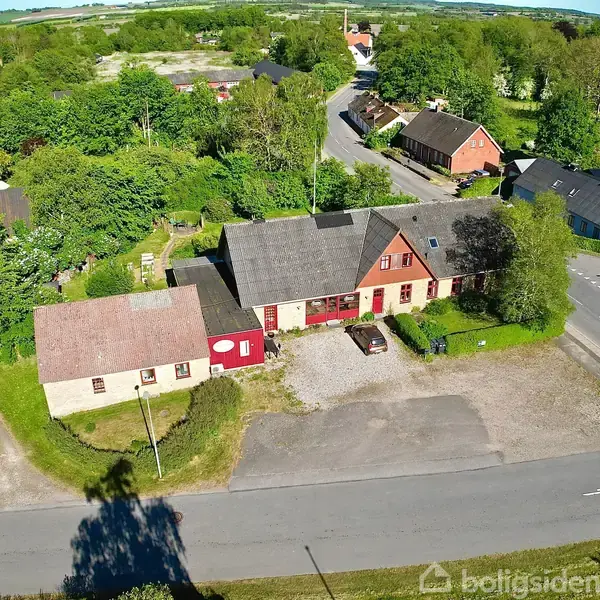 En række gamle bygninger ligger langs en landevej i et grønt, skovklædt område. Bygningerne har forskellige røde og beige farver med gråt tag, og der er en gårdsplads foran.