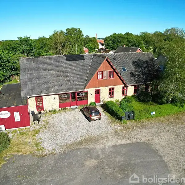 Et hus med rødmalede vinduer og døre står ved en grusparkering. Foran huset holder en bil parkeret. Omgivelserne inkluderer træer og en vej og navnet "Humlehuset".