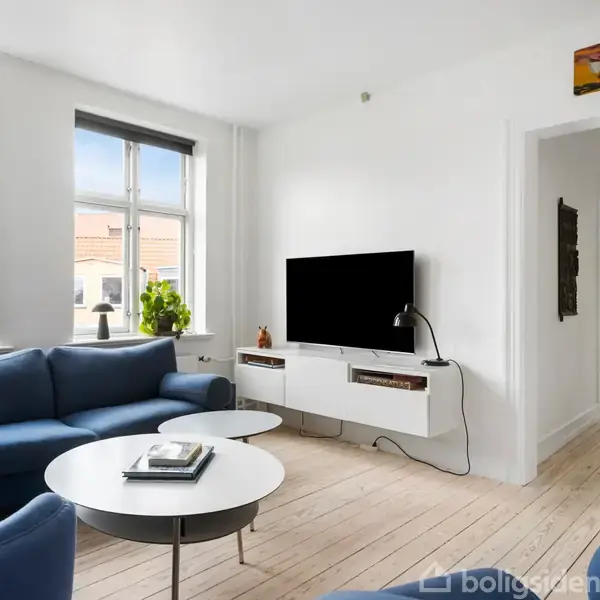 En stue med blå sofaarrangement omgiver to runde borde. Et TV er monteret på væggen over en hvid skænk. Rummet har store vinduer med udsigt til tage.