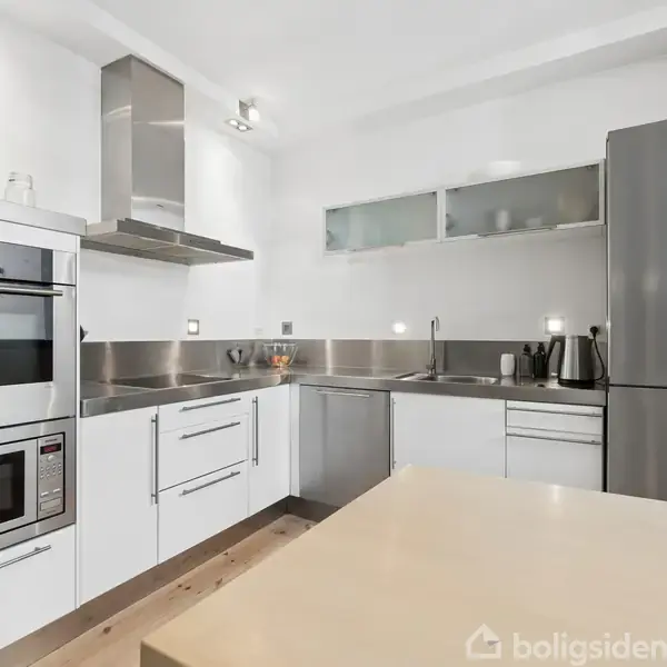 Et moderne køkken med hvide skabe, rustfri metalbordplader og en integreret ovn. Det indeholder også en stålkøleskab ved siden af en elkedel og toaster. Flaske og krydderier pryder hylden.
