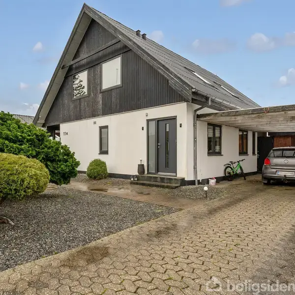 Et hus med sort tag og hvid facade står i en have med grå belægning og grøn beplantning. En bil er parkeret i en carport til højre.