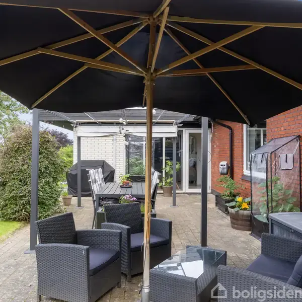 Parasol dækker havemøbler i flet på en brolagt terrasse foran et rødt murstenshus. Planter i potter og en grill ses omkring, med en grøn have i baggrunden.