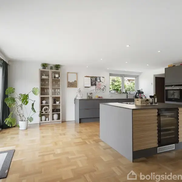 Et moderne køkken med en stor kogeø i midten med indbygget vinkøleskab. Køkkenet har lyse træ- og grå overflader. Store vinduer til venstre giver naturligt lys og kig til haven.