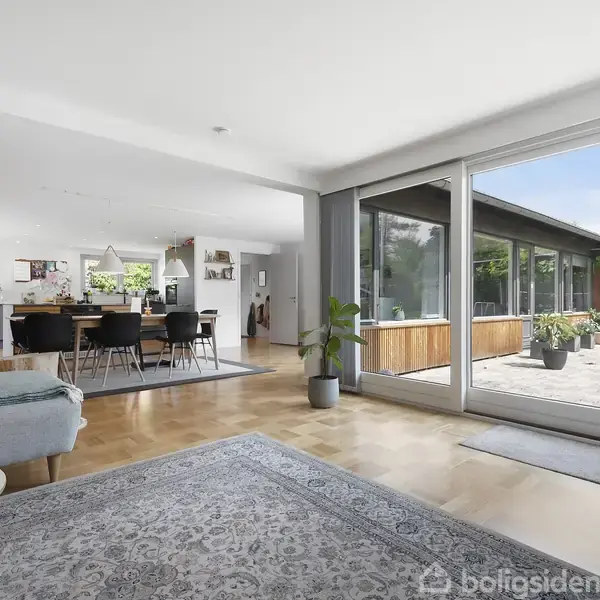 Stue med sofa og tæppe, der åbner mod en terrasse med glasdør. I baggrunden et spisebord i et lyst og moderne rum med trægulve og grønne planter.