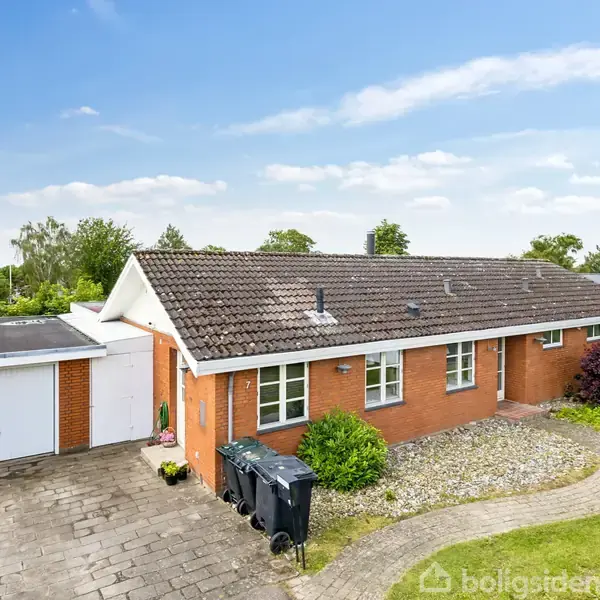 En étplans, rød murstensvilla med mørkt tag ligger i en velholdt have. Der er en flisebelagt indkørsel og en tilstødende garage. Hække og græsplæne omkranser huset.