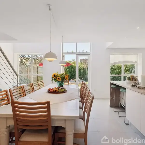 Et lyst køkken-alrum med et langt spisebord og træstole. Bordet er dekoreret med en vase med solsikker og en skål med frugt. Omkring er åbne vinduer og et moderne køkken med en ø.