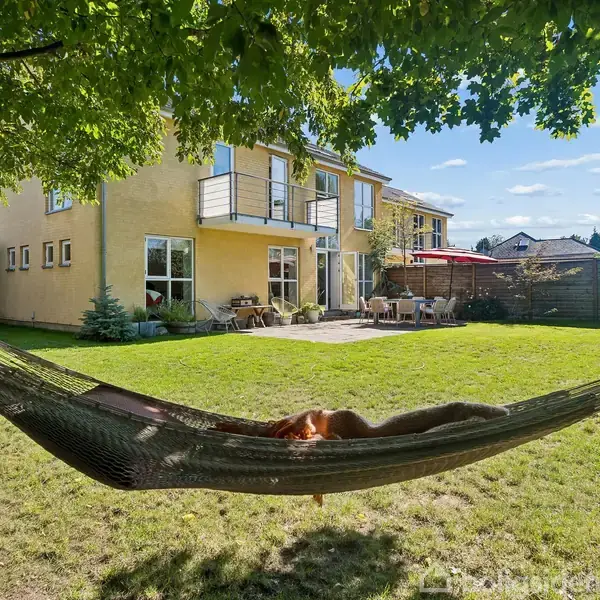 En hængekøje hænger i skyggen af et træ, hvor nogen slapper af. I baggrunden ses et gult hus med balkon og terrassemøbler, omgivet af græsplæne og buske.