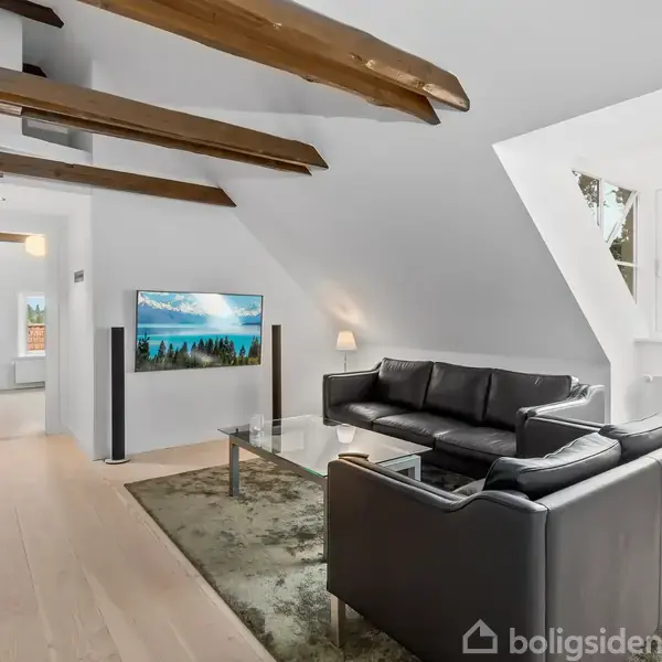 Sort læderhjørnesofa står på et tæppe foran et tv på væggen; placeret i en stue med skråt loft, synlige bjælker og lyse trægulve.