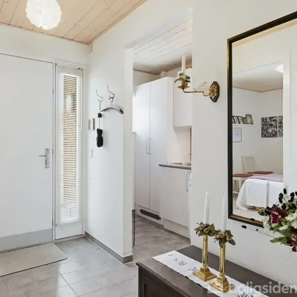 Indgangsparti med en hoveddør og smalt vindue. En skænk med blomster og lysestager. En vægdekoreret lampe og et spejl. Køkken og stue kan skimtes i baggrunden.