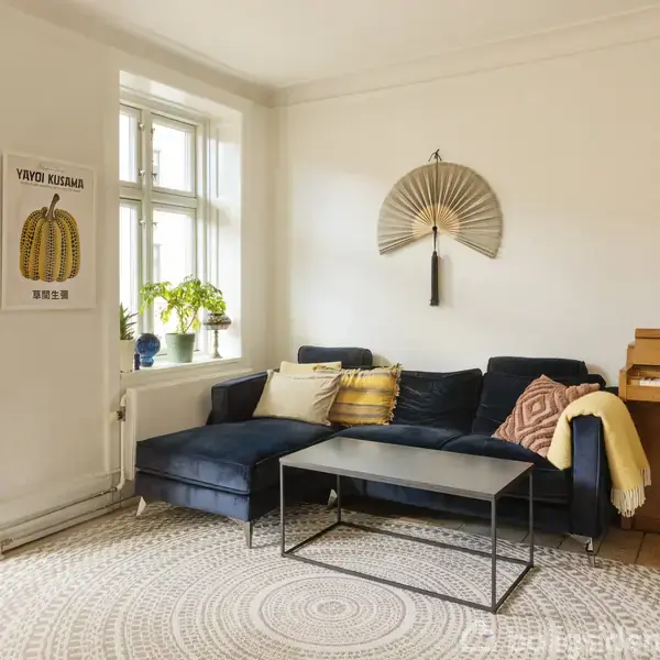 En blå hjørnesofa med puder står i en hyggelig stue, hvor en sort bord placeret foran. Væggen har en stor vifte og en guitar. Et klaver står ved siden af sofaen. En plakat med "YAYOI KUSAMA" hænger ved vinduet.