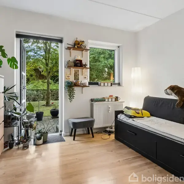 En stue viser en sofa med en brun pude, der hviler på en gråmadras. Ved siden af er der en kommode. Et åbent vindue fører til en terrasse med planter.