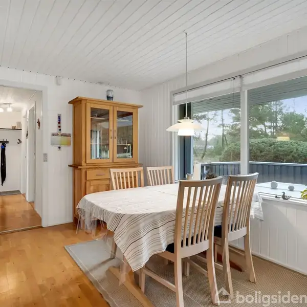 Spisebord dækket med en dug og omgivet af træstole står i lys spisestue. Vindue bagved viser buske udenfor. Et vitrineskab ses i baggrunden. Gulvet er af træ.