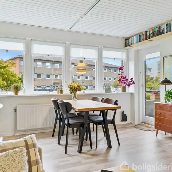 Spisebord med stole står i en stue med store vinduer, som giver udsigt til boligblokke. Rummet er lyst, med planter og hylder fyldt med bøger.