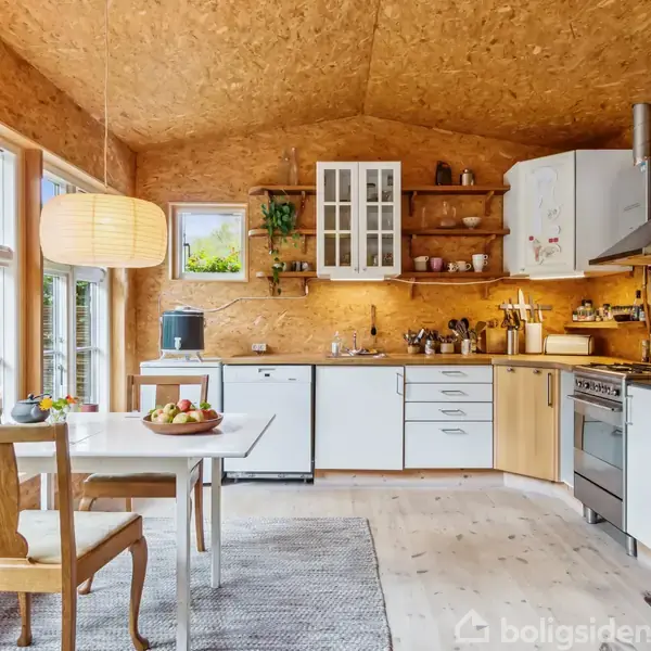 Køkken med trævægge og lofter, indeholder hvidt køkkenskab, gasovn og spisebord. Stor vindue mod venstre, bringer naturligt lys ind. Hylder med planter og køkkenartikler pynter væggene.