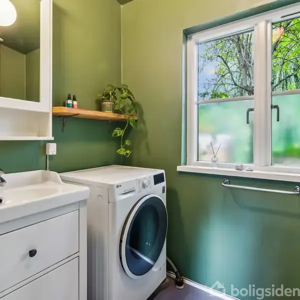 En vaskemaskine står ved siden af en hvid vask i et grønt badeværelse med et vindue, der viser træer udenfor. Håndvasken har en sæbedispenser, og der er en hylde med planter.