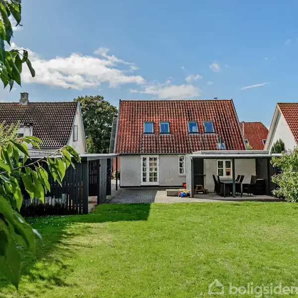 Hus med rødt tag og flere vinduer står i centrum, omgivet af en grøn have med træer. En carport er til venstre, og en terrasse med borde er til højre.