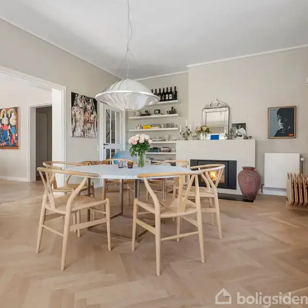 Et rundt spisebord med træstole står i et lyst rum. Over bordet hænger en moderne lampe. Rummet er dekoreret med kunstværker og har en pejs og hylder med flasker.