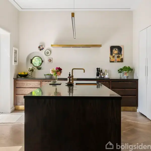 Køkkenø med sort bordplade står centralt, omgivet af træskabe. Dekorationer, blomster og en vase pryder bordet, mens moderne kunst hænger på de lyse vægge. Lysindfald fra venstre.