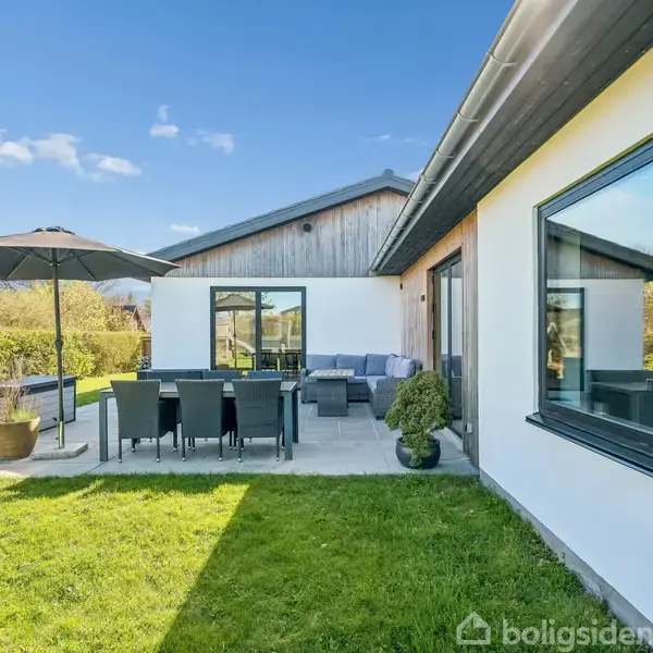 Hus med hvide mure og sort tag udendørs. Terrasse med spisebord under parasol, omgivet af græsplæne. Blåt himmel med spredte skyer. Vegetation i baggrunden.