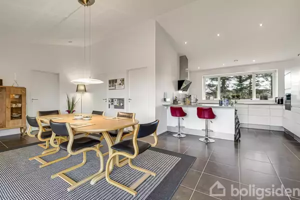 Rundt spisebord med træstole står på et tæppe i et moderne køkken-alrum, hvor to røde barstole er placeret ved en køkkenø. Store vinduer giver udsigt til træer udenfor.